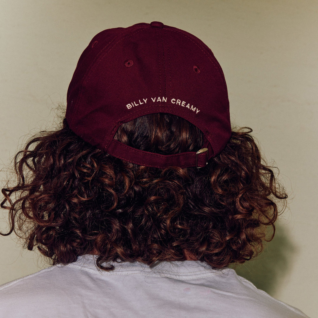 a burgundy red cap viewed on a person with curly hair from behind. The embroidery on the back of the cap says 'BILLY VAN CREAMY'